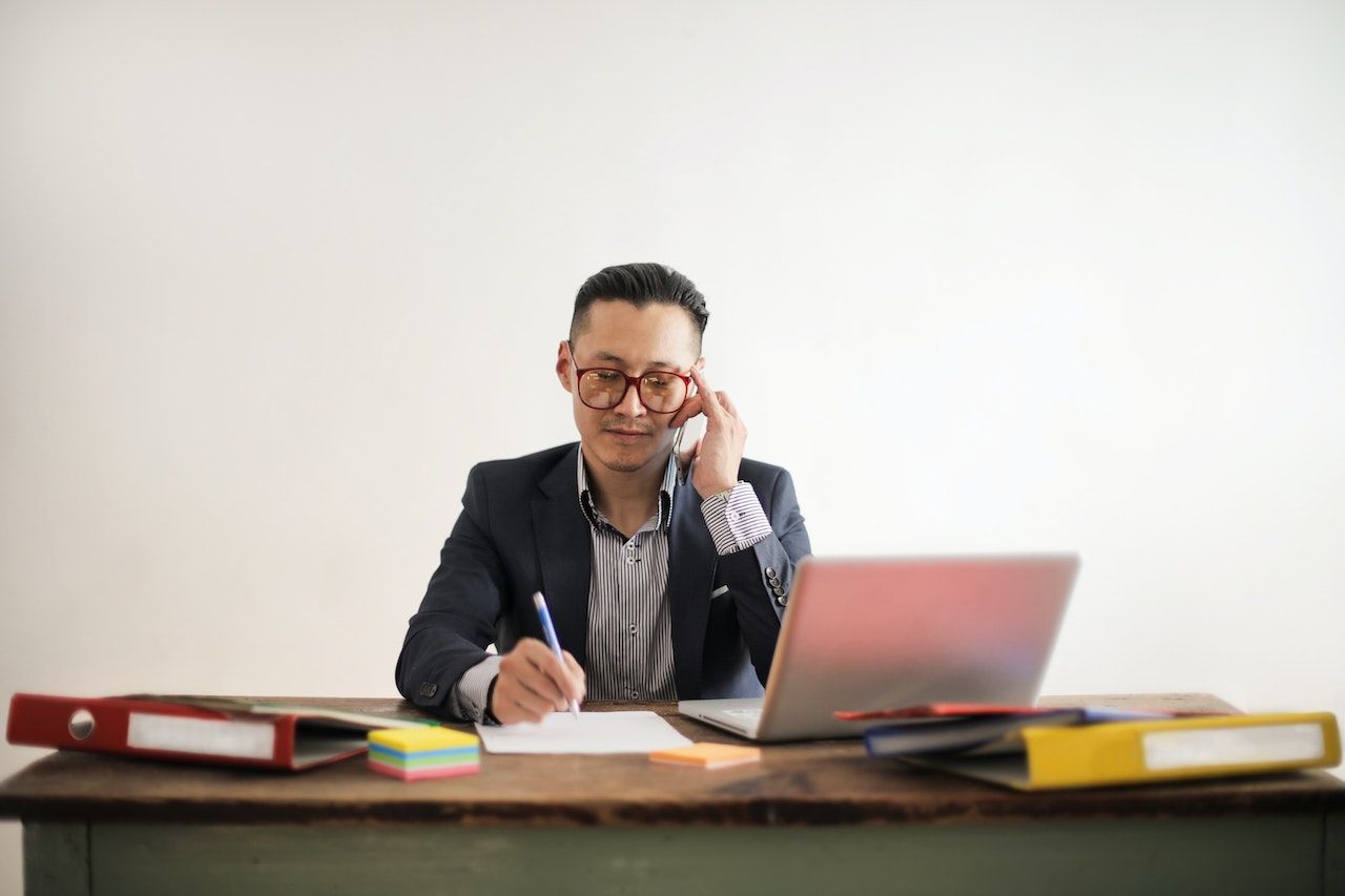 man in blue suit and eyeglasses talking on the phone while sitting at a table with his laptop